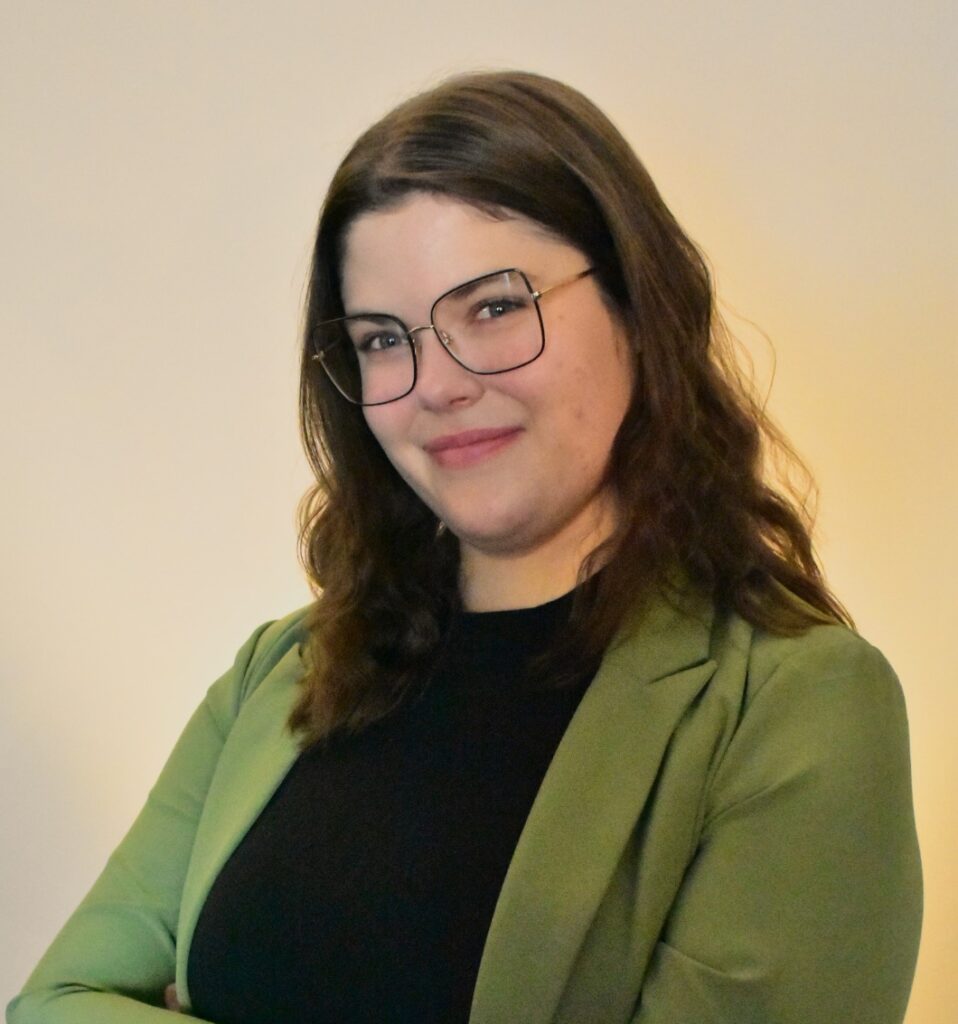 Young woman wearing a green blazer over a black shirt with eye glasses and brown hair.